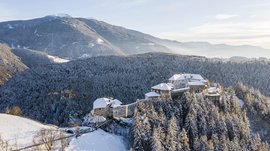 Rodeneck – for a wonderful walking holiday in Italy The picture shows a castle located on a wooded hill covered with snow. The castle itself has snow-covered roofs and is surrounded by dense forests. In the background, gentle hills and mountains stretch out under a clear, blue sky.