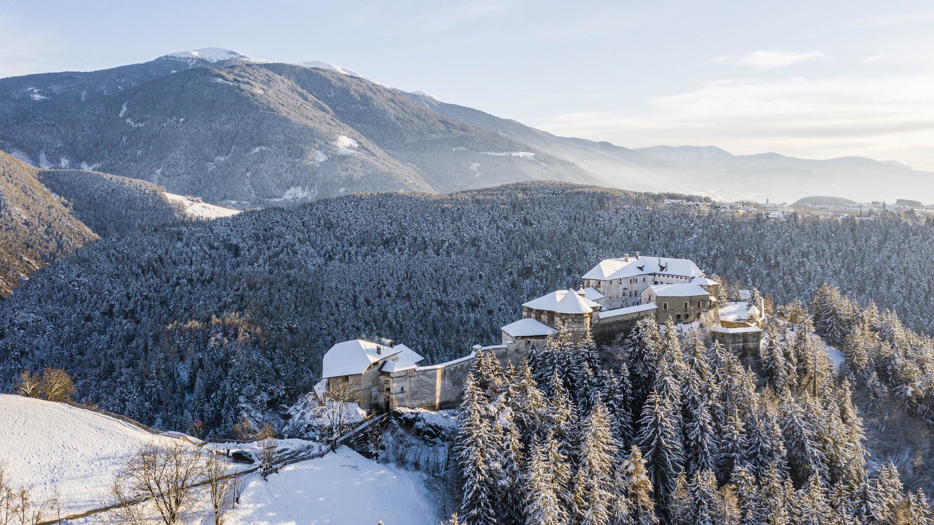 Rodeneck – for a wonderful walking holiday in Italy The picture shows a castle located on a wooded hill covered with snow. The castle itself has snow-covered roofs and is surrounded by dense forests. In the background, gentle hills and mountains stretch out under a clear, blue sky.