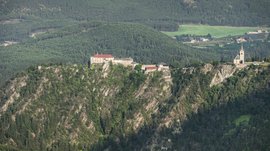 Rodeneck – for a wonderful walking holiday in Italy The picture shows a castle and a church perched on a steep rock. Both buildings are surrounded by lush, green forest that stretches over the surrounding hills.