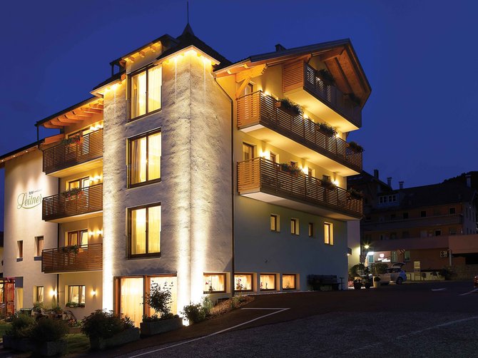 Willkommen in der Ferienregion Gitschberg Jochtal! Das Bild zeigt ein beleuchtetes, mehrstöckiges Hotelgebäude bei Nacht. Die Fassade des Gebäudes ist gelblich beleuchtet, und auf den Balkonen befinden sich Blumenkästen. Das Gebäude strahlt eine warme, einladende Atmosphäre aus, während im Hintergrund ein dunkler Nachthimmel zu sehen ist.