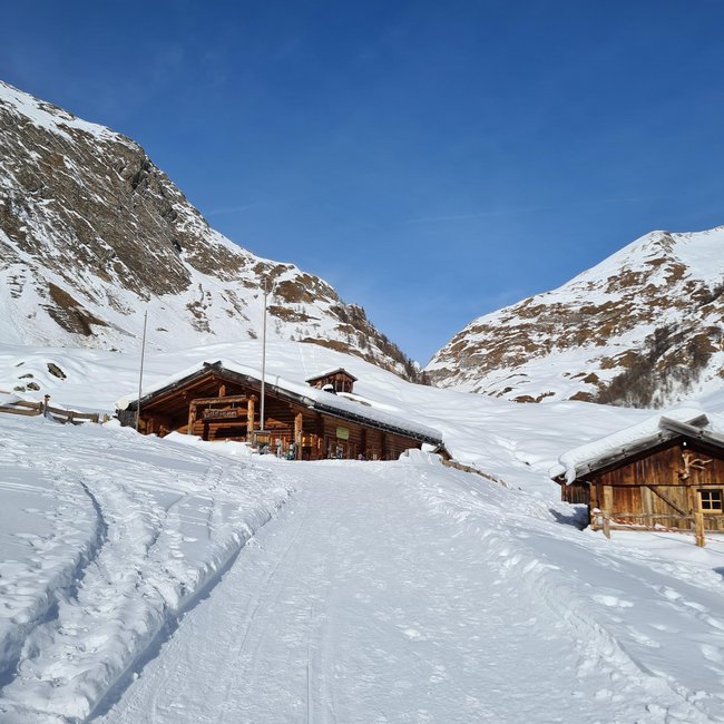 Kuttnhütte Das Bild zeigt eine Berghütte aus Holz, umgeben von tiefem Schnee, auf einem verschneiten Hang. Im Hintergrund ragen schneebedeckte, felsige Berge unter einem klaren, blauen Himmel auf.