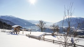 Rodeneck – for a wonderful walking holiday in Italy The picture shows a snowy landscape with a wooden fence, a small wooden cabin, and trees in the foreground. In the background, a vast valley stretches out, surrounded by tall snow-covered mountains, while the sun shines brightly in the blue sky.