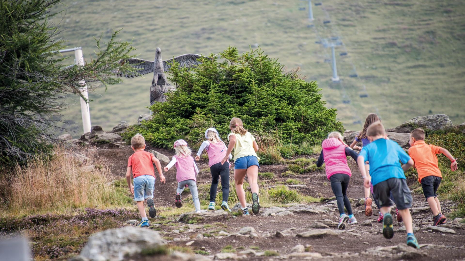 Sonnenpark Gitschberg De afbeelding toont een groep kinderen die een rotsachtig pad oplopen. Op de achtergrond is een groot beeld van een adelaar met uitgespreide vleugels te zien, verborgen achter een struik.
