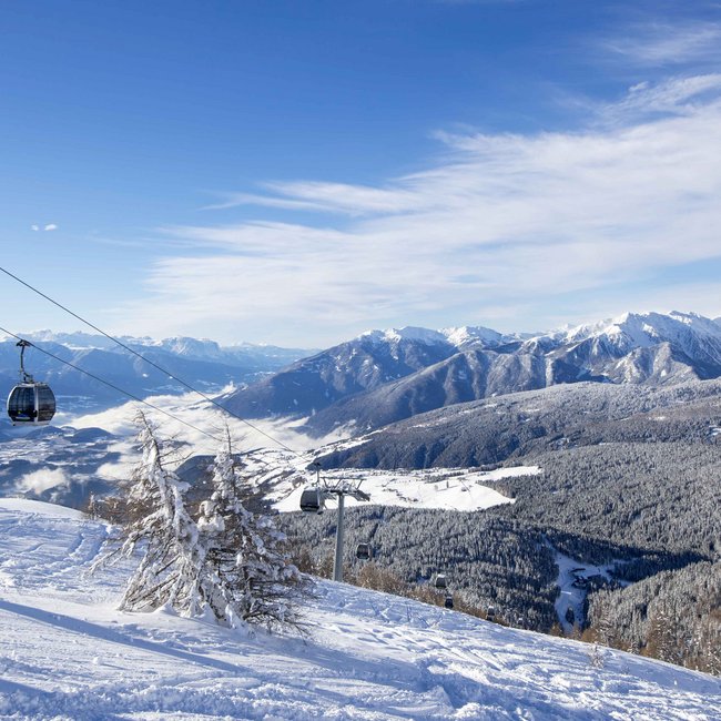Find your hotel near Brixen! Cable car cabins over snowy mountains and trees under a blue sky