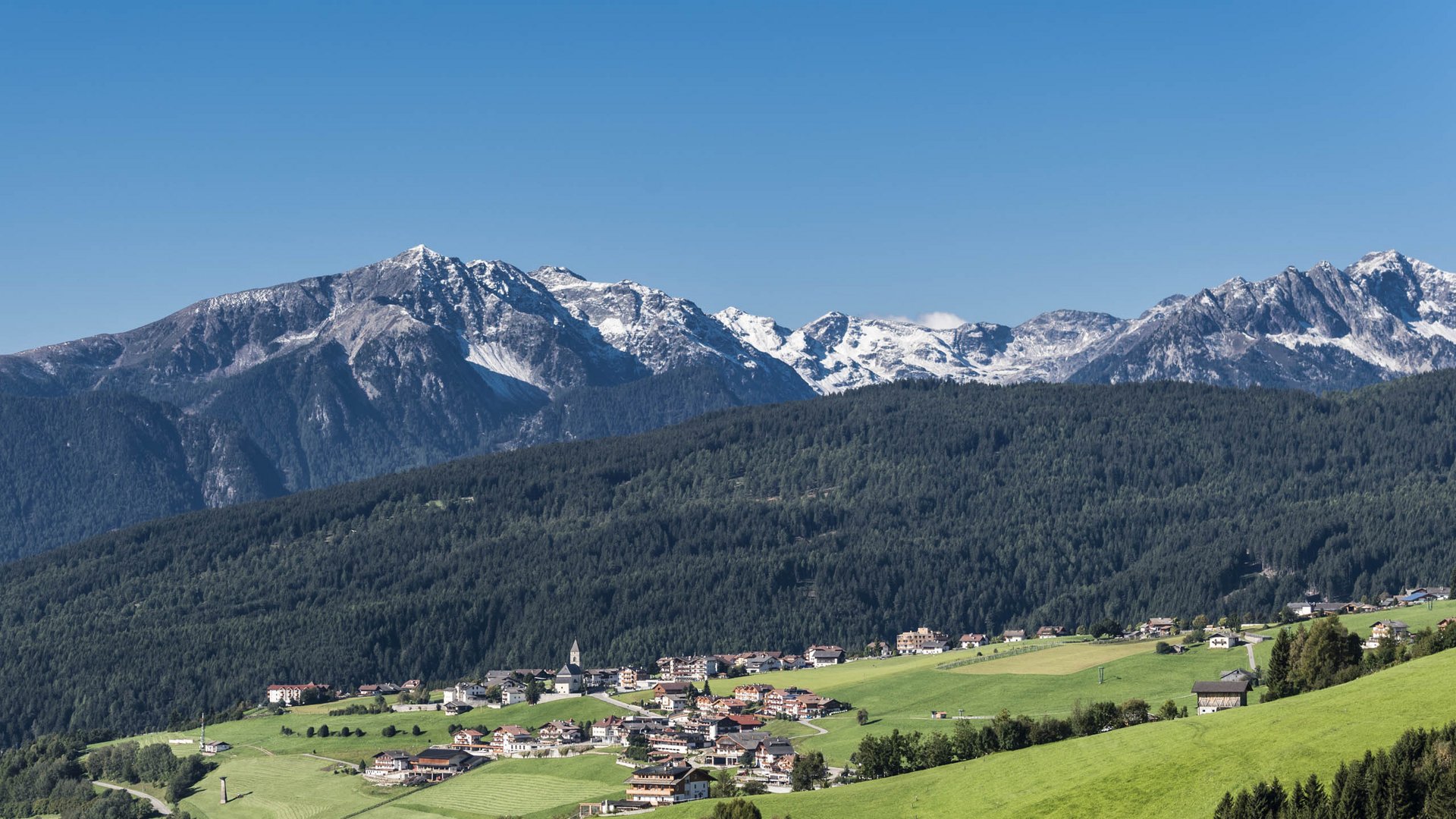 Das Bergdorf Meransen Das Bild zeigt ein Dorf, das sich über sanft abfallende grüne Hügel erstreckt, vor einem Hintergrund dichter Wälder und schneebedeckter Berge. In der Mitte des Dorfs steht eine Kirche mit einem spitzen Turm, umgeben von Häusern, und die gesamte Szene wird von einem klaren blauen Himmel überdacht.