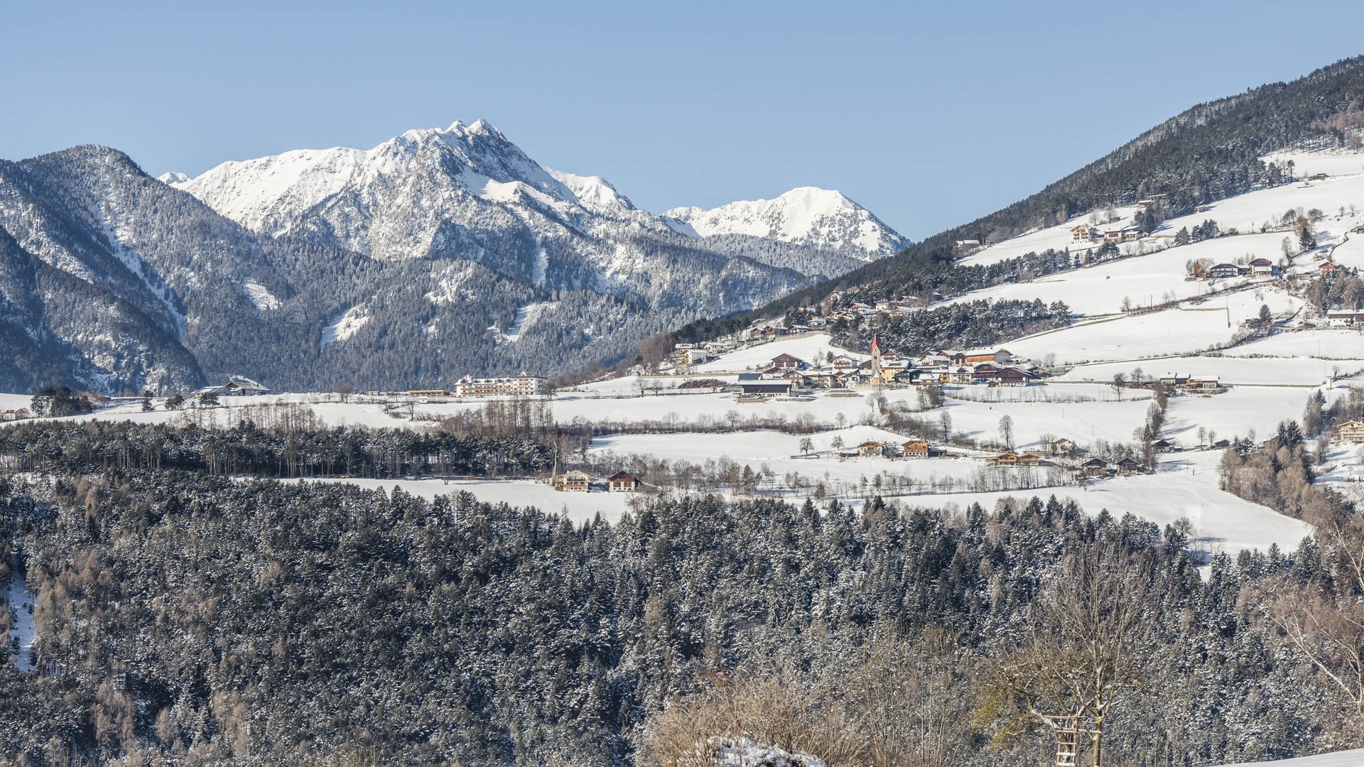 The Alpine hamlet of Spinges The picture shows a snowy landscape with a small village in the foreground, surrounded by snow-covered fields and forests. In the background, impressive mountains rise with snow-covered peaks under a clear blue sky.