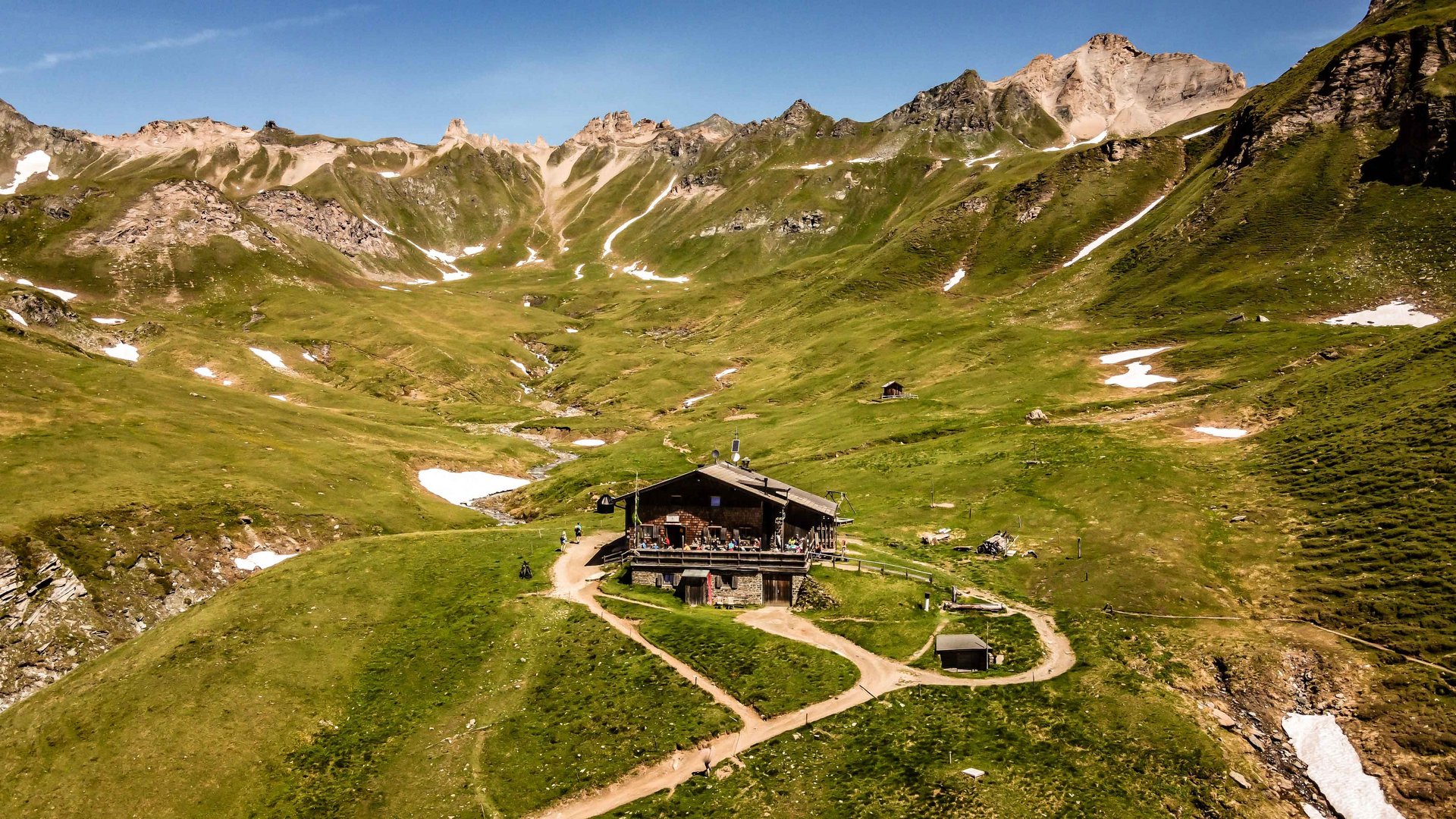 The Pfunders High Mountain trail The picture shows an alpine hut in the middle of a green mountain landscape with scattered patches of snow. In the background, steep, rocky mountains rise under a clear blue sky. A small stream flows through the valley beside the hut.