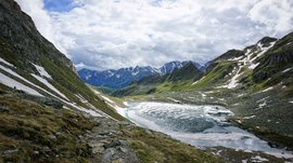 The Pfunders High Mountain trail The picture shows a mountain landscape with snow-covered peaks in the background. A partially frozen lake lies in a valley, surrounded by rocky paths and grass-covered slopes.