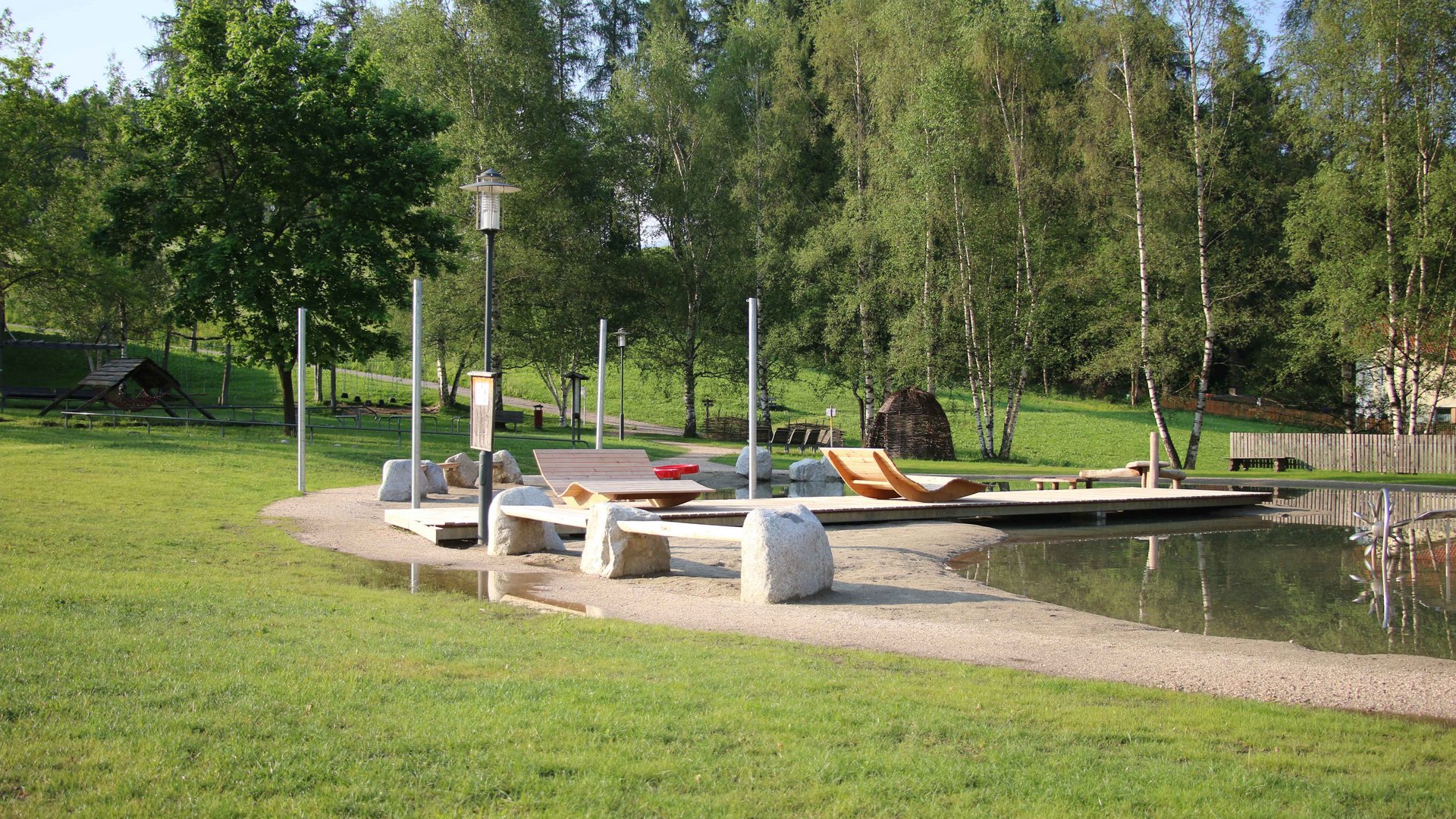 Aktivpark Terenten Obrázek ukazuje klidný park s malým rybníkem, ve kterém se nacházejí dřevěná lehátka na písčité ploše. V pozadí je několik stromů a okolí je plné zeleně a přírody.