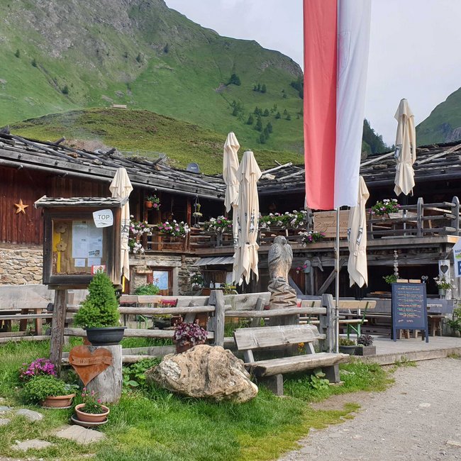 Zingerle Hütte Obrázek zobrazuje rustikální alpskou chatu s dřevěnými a kamennými stěnami, obklopenou zelenými kopci. Před chatou stojí několik dřevěných lavic a stolů, některé s uzavřenými slunečníky, a je zde mnoho barevných květin a dřevěná socha. V popředí vlaje velká červeno-bílá vlajka.