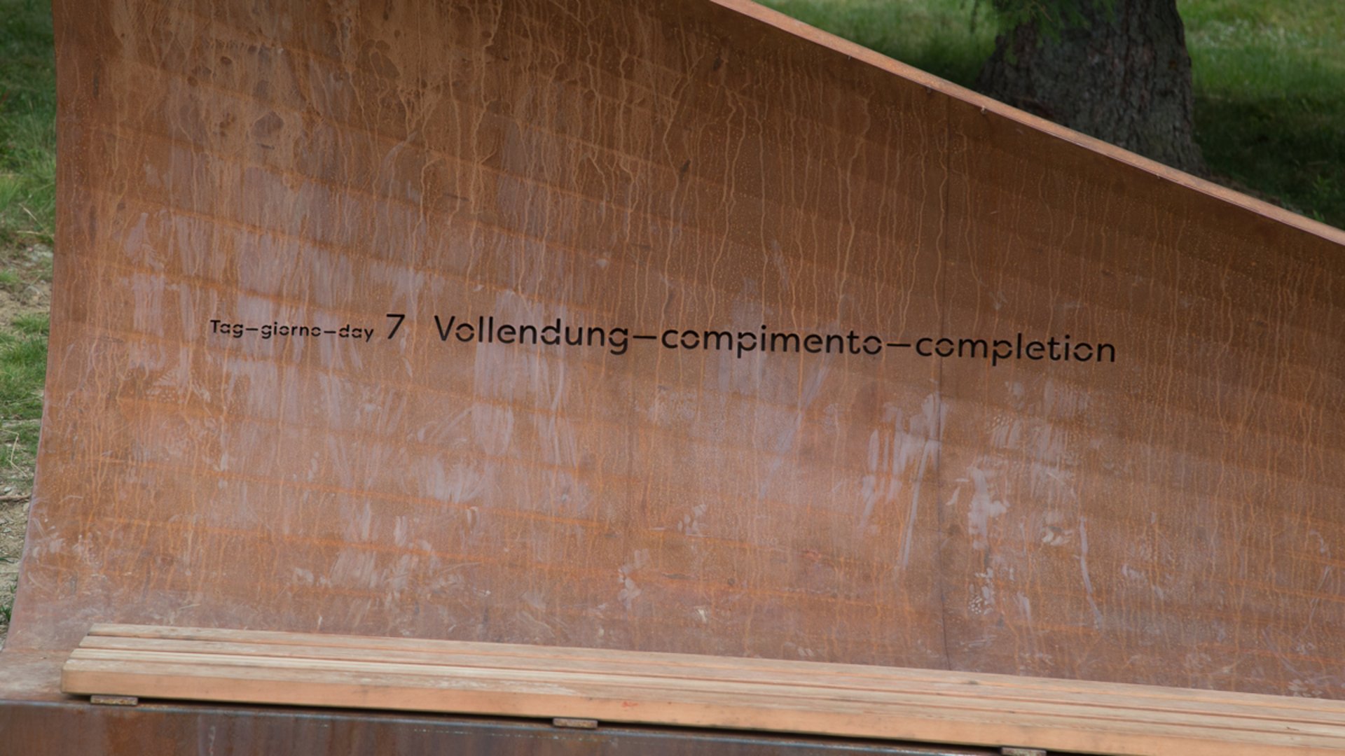 The Creation trail in Rodeneck The picture shows a metal bench or sculpture, on which the inscription "Tag 7 – Vollendung" is engraved in several languages. The structure appears rust-colored and stands outdoors, presumably in an unspoiled natural environment.