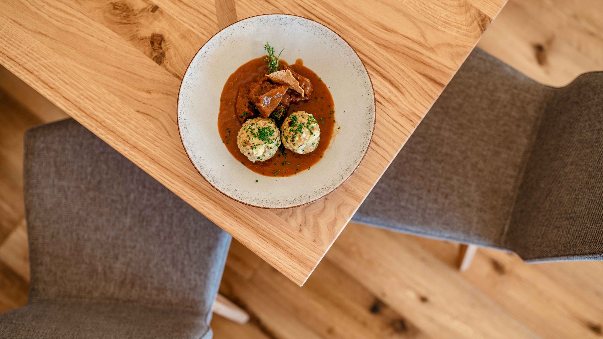 Discover the Alpine huts in Meransen, Vals & Co. The picture shows a modernly presented plate with two bacon dumplings and goulash on a wooden table, seen from a bird's eye view, with two empty chairs in the background.