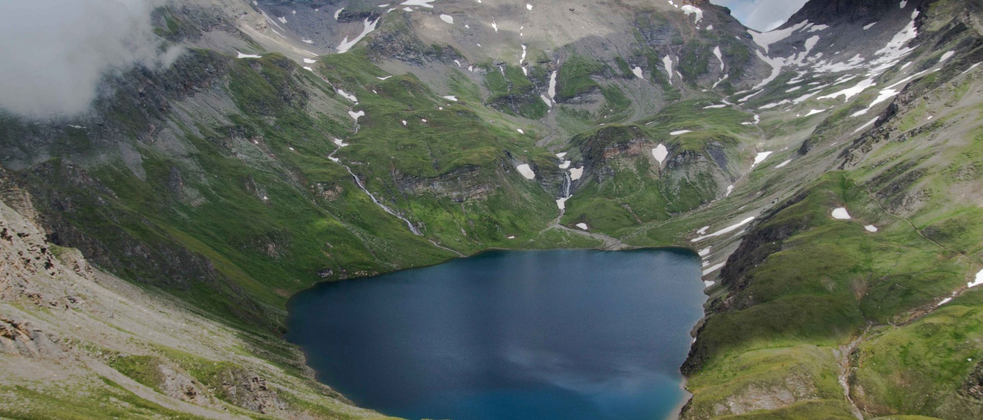 Wilde Kreuzspitze Das Bild zeigt einen hochgelegenen Bergsee, umgeben von steilen, grünen Berghängen und schneebedeckten Gipfeln. Der Himmel ist wolkenverhangen, was der Szenerie eine dramatische Atmosphäre verleiht.
