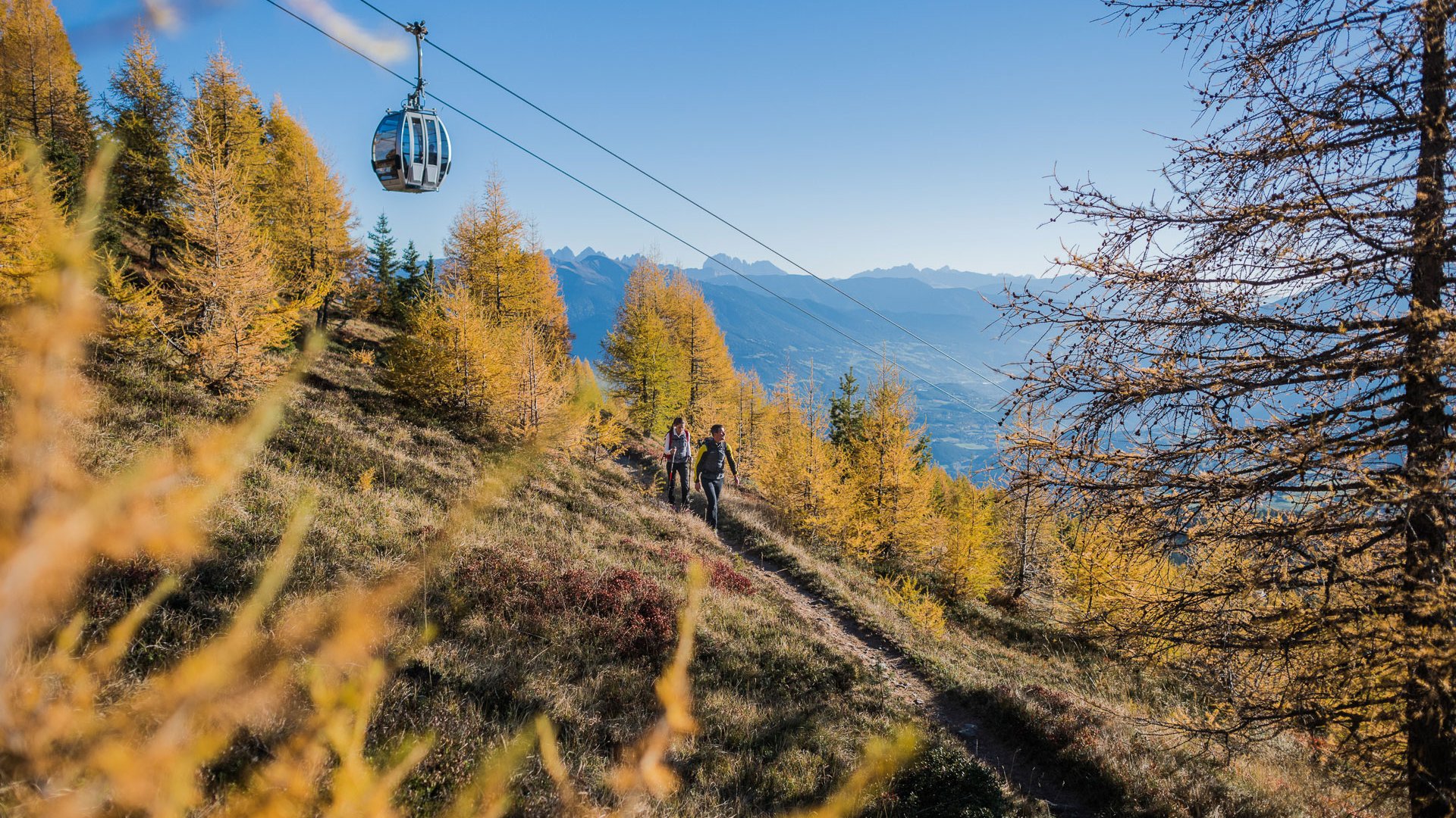 Willkommen in der Ferienregion Gitschberg Jochtal! Das Bild zeigt eine Seilbahn, die über einen Pfad fährt, der von Wanderern benutzt wird. Die Landschaft ist herbstlich, mit goldenen Bäumen, und im Hintergrund sind Berge unter einem klaren blauen Himmel zu sehen.