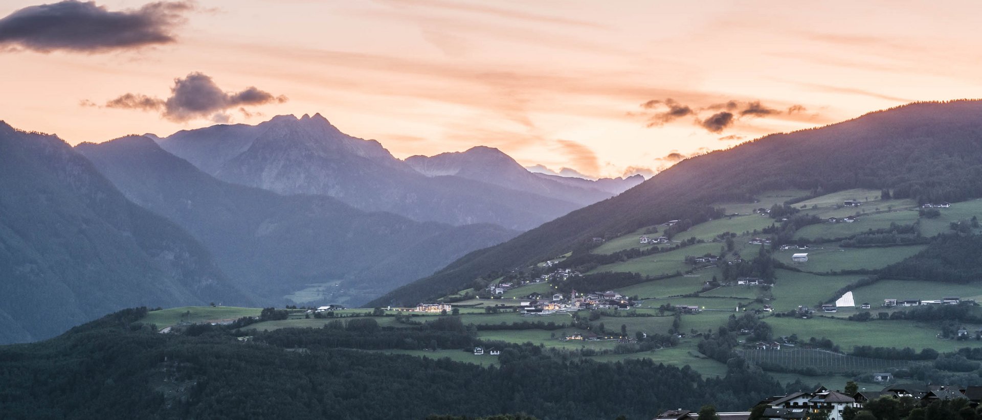 Willkommen in der Ferienregion Gitschberg Jochtal! Das Bild zeigt eine friedliche Berglandschaft bei Sonnenuntergang. Die Berge sind in dunklen Silhouetten zu sehen, während der Himmel in sanften Orangetönen leuchtet. Im Vordergrund erstrecken sich grüne Hügel mit verstreuten Häusern.